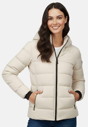 Woman with dark hair smiles while wearing a light beige puffer jacket with a hood and hands in zippered pockets, paired with blue jeans.