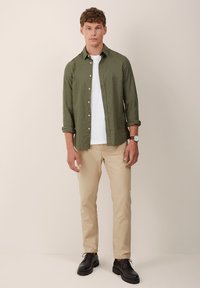 Cămașă verde cu nasturi peste un tricou alb, pantaloni bej și pantofi negri. Croială ajustată, țesătură ușoară, cu manșete de mânecă răsfrânte și un ceas de mână ca accent.