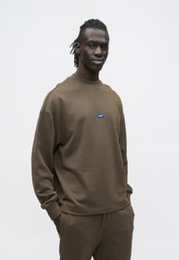 Felpa marrone con collo a costine, maniche lunghe e logo rettangolare blu sul petto. Presenta una vestibilità comoda e una texture morbida.