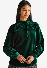 Samtgrüne Bluse mit hohem Rüschkragen, langen Puffärmeln und einer vorderen Öffnung, die durch eine Rüsche betont wird. Weicher Stoff und elegantes Design.