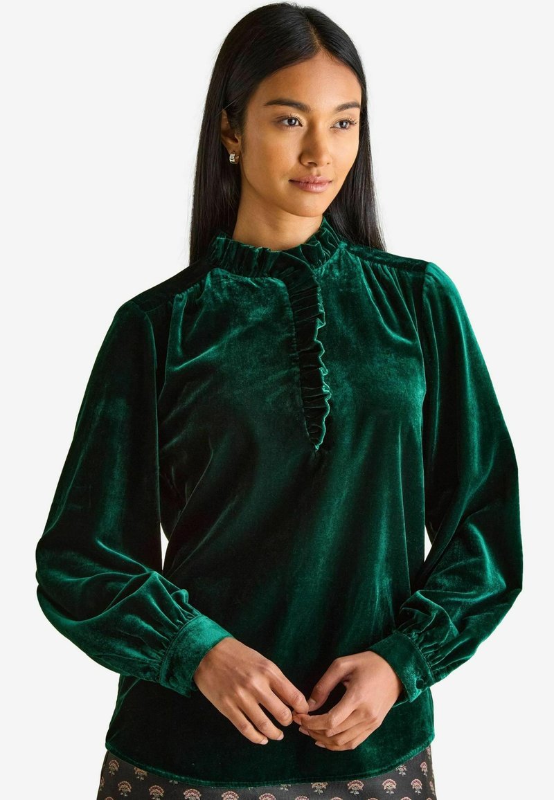 Samtgrüne Bluse mit hohem Rüschkragen, langen Puffärmeln und einer vorderen Öffnung, die durch eine Rüsche betont wird. Weicher Stoff und elegantes Design.