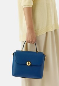 Mano che tiene una borsa in pelle blu con chiusura dorata, indossando un maglione crema e una gonna beige.
