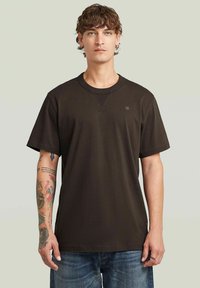 T-shirt in cotone marrone scuro con collo rotondo, maniche corte e un piccolo logo sul petto. Vestibilità ampia con una texture liscia.
