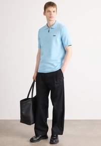 Jovem vestido com uma polo de mangas curtas azul claro, calças largas pretas, mocassins pretos, segurando uma grande bolsa tote preta.