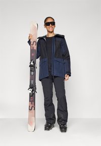 Skiausrüstung umfasst eine navy- und schwarzfarbene wasserdichte Jacke, schwarze Hose und schwarze Skischuhe. Das Snowboard hat ein grau-verlaufendes Design und Bindungen.