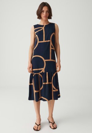 Donna con capelli corti che indossa un abito blu navy senza maniche con motivi geometrici beige e sandali neri con tacco, in piedi su uno sfondo semplice.