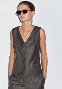 Kobieta w ciemnej kamizelce w paski i pasujących spodniach bez rękawów, z rękami w kieszeniach i prostokątnych ciemnych okularach przeciwsłonecznych.