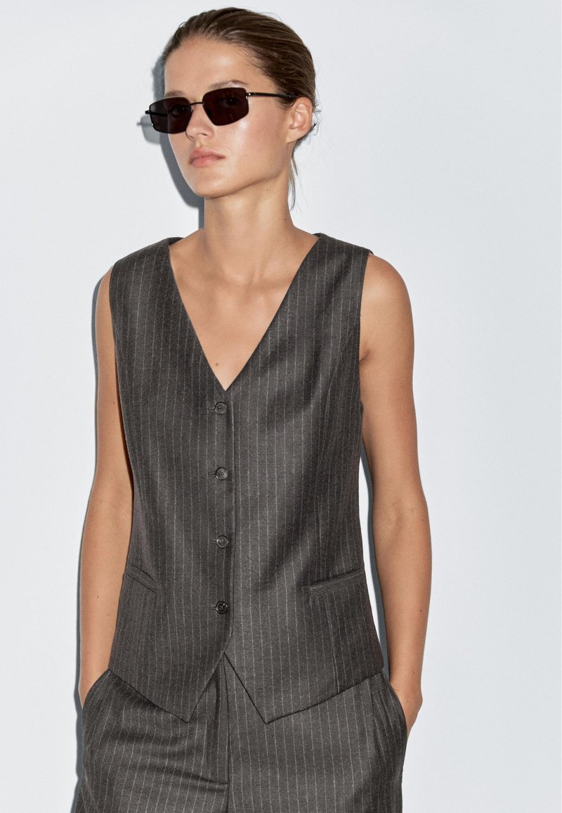 Kobieta w ciemnej kamizelce w paski i pasujących spodniach bez rękawów, z rękami w kieszeniach i prostokątnych ciemnych okularach przeciwsłonecznych.