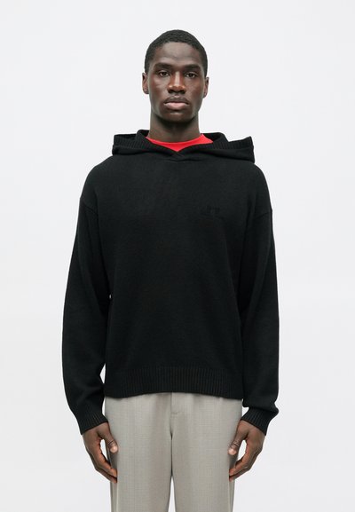 Homme portant un sweat à capuche noir sur une chemise rouge et un pantalon gris clair, se tenant devant un fond blanc uni.