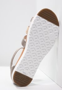 Biała gumowa podeszwa damskich sandałów z brązowym obcasem i uchwytami na palce, metaliczne paski oraz znak "Treadlite by UGG" w pobliżu pięty.