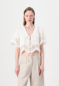 Hvit kortermet bluse med bølget kant, utskårne blomsterdesign, korte ermer og knytelukking foran. Kombinert med lysebeige bukser.