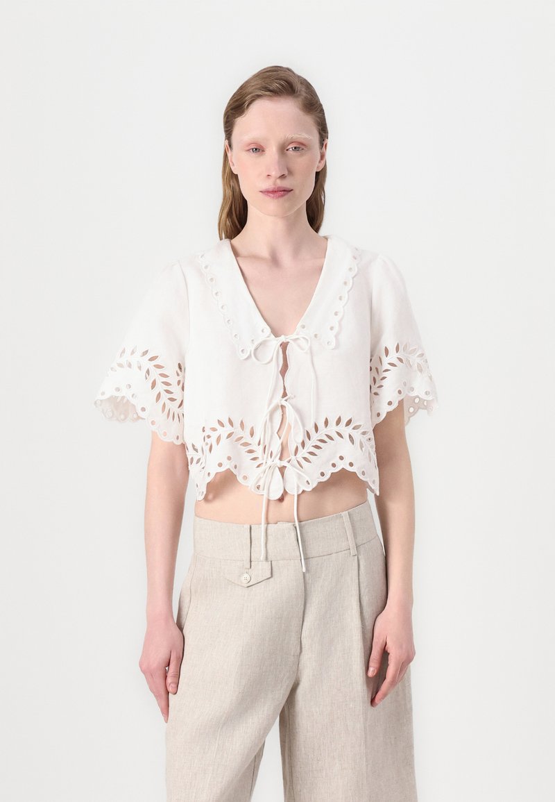 Hvit kortermet bluse med bølget kant, utskårne blomsterdesign, korte ermer og knytelukking foran. Kombinert med lysebeige bukser.