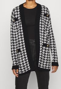 Zwart-wit hakketakk cardigan met een ribbelkraag in het zwart, open voorkant en twee nepzakken met gouden knopen. Zachte gebreide textuur.