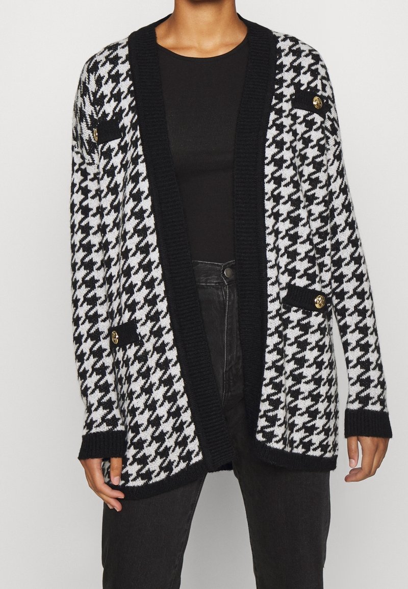 Zwart-wit hakketakk cardigan met een ribbelkraag in het zwart, open voorkant en twee nepzakken met gouden knopen. Zachte gebreide textuur.
