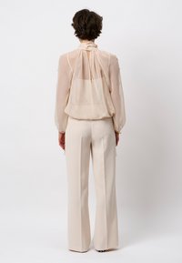 Helle beige, durchsichtige Bluse mit hohem Kragen und gerafftem Rücken, kombiniert mit locker sitzenden, cremefarbenen Hosen. Glatte Textur, elegante Silhouette.