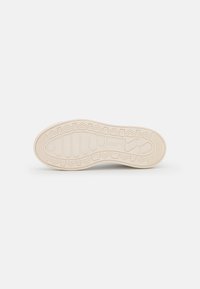 Shoe outsole in off-white rubber, featuring a patterned tread with geometric shapes and logo detail. Smooth texture with varying thickness.