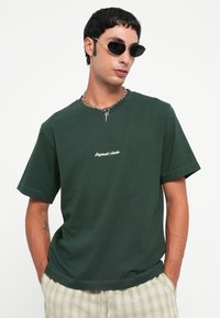 Bărbat îmbrăcat cu un tricou „Originals Studio” de culoare verde închis, cu o lanț argintie la gât, ochelari de soare negri, ovali și pantaloni deschiși la culoare, cu un model în carouri, cu mâinile în buzunare.