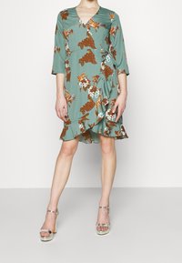 V-kauluksellinen teal-värinen mekko, jossa on ruskeita ja sinisiä kukkakuoseja, kolme neljänneksen hihat ja röyhelöhelma. Yhdistettynä hopeisiin korkokenkiin.