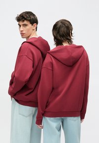 Bordó kapucnis pulóver laza szabással, bordázott mandzsettákkal és zsebbel. Világoskék farmernadrággal viselve, elölről és hátulról nézve.