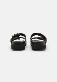 Schwarze Sandalen mit flacher Sohle, die mehrere Riemen und ein silbernes Schnallendetail aufweisen. Die Textur wirkt glatt und hat ein minimalistisches Design.