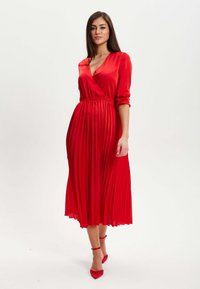 Red, pleated midi dress with long sleeves and a deep V-neck. Smooth satin material with an elastic waistband and matching red heels.