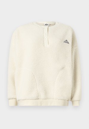 Pull en polaire couleur crème avec des manches longues, patte de boutonnage au col, poignets et ourlet côtelés, et petit logo triangulaire sur la poitrine.