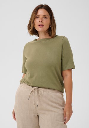 Femme aux cheveux bruns mi-longs portant un haut vert olive à manches courtes et un pantalon beige à cordon, debout devant un fond clair uni.