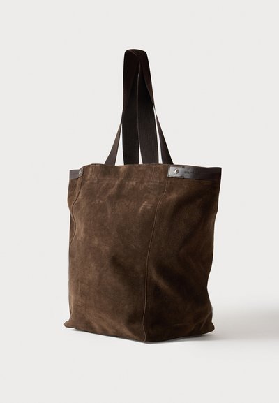 Sac cabas en daim marron avec anses en tissu foncé et détail de garniture en cuir sur un fond clair.