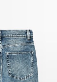 Jean en denim bleu clair avec une taille mi-haute, des coutures visibles et une seule poche arrière. Le tissu présente une texture légèrement délavée.