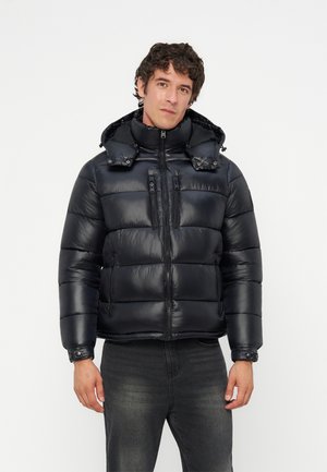 Homme portant une veste matelassée noire avec une capuche, fermée par une fermeture éclair, associée à un jean gris foncé, debout devant un fond blanc uni.