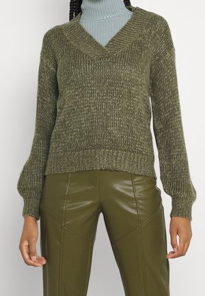 Pull en maille vert olive avec un décolleté en V, des poignets et un ourlet côtelés. Associé à un pantalon en cuir verni vert olive ajusté.