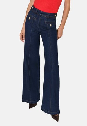 Kvinde iført højtaljede, mørkeblå brede jeans med knapdetaljer i taljen og en rød ærmeløs top, stående mod en hvid baggrund.
