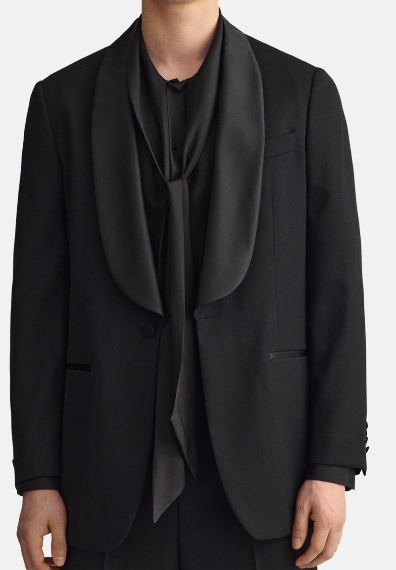 Giacca da smoking nera con colletto a scialle in satin, abbinata a una sciarpa nera coordinata drappeggiata attorno al collo, texture liscia, vestibilità sartoriale.