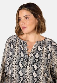 Blouse à manches longues en imprimé serpent, présentant des tons neutres de noir et beige, avec un décolleté en V et des accents de boutons métalliques.