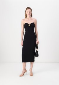 Čierne midi šaty bez ramienok so srdcovým výstrihom a akcentom zlatého kovu. Modelka drží malú čiernu kabelku a má na sebe telové lodičky.