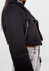 Svart kort jacka med en fluffig krage, silverdragkedjedetalj och strukturerad tyg. Har en sidodragkedjeficka och släta ärmar.