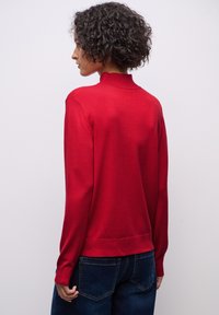 Roter Strickpullover mit hohem Kragen, gerippten Bündchen und Saum, kombiniert mit dunklen Jeans. Glatte Textur, figurbetonter Schnitt und einfarbig.