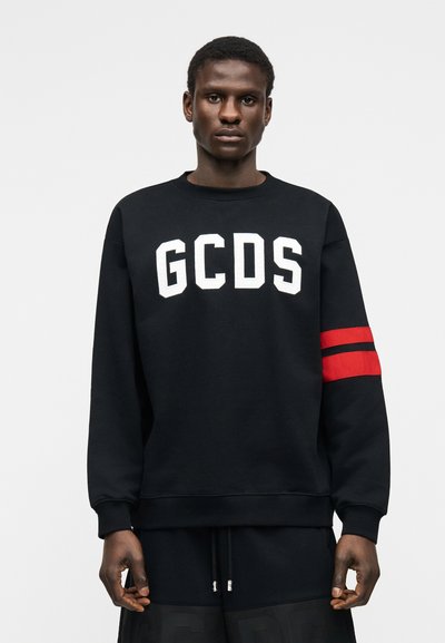 Νέος άνδρας που φοράει μαύρο φούτερ με λευκά γράμματα "GCDS" και κόκκινες και μαύρες ρίγες στο αριστερό μανίκι, στέκεται μπροστά σε ένα απλό φόντο.