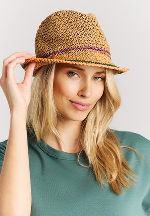 Blonde vrouw in een groen shirt past een gevlochten strohoed met paarse en groene strepen aan tegen een lichtgrijze achtergrond, met een subtiele glimlach.