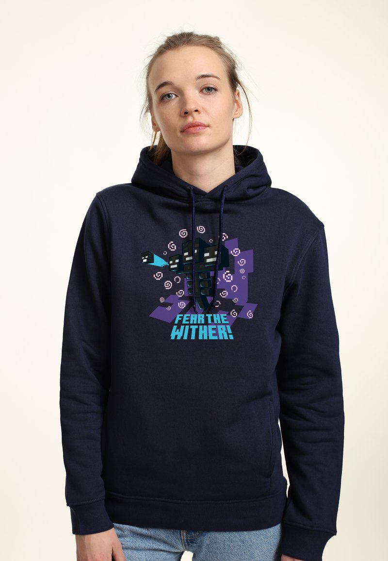 Giovane donna che indossa un hoodie blu scuro con una creatura a tre teste pixelata e il testo "Temi il Wither!" su uno sfondo chiaro.