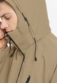 Beige wasserdichte Jacke mit hohem Kragen, ausgestattet mit einer Kordelzugkapuze und schwarzen Reißverschlussdetails. Glattes Material ohne Muster oder Akzente.