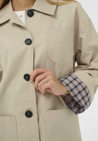 Manteau beige boutonné avec des boutons noirs et une poche poitrine, porté sur une chemise à manches retroussées à carreaux, montrant une main qui ferme un bouton.