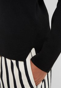 Sweater noir en tricot avec une forme d'ourlet en V, associé à un tissu rayé en blanc et bleu marine. Gros plan sur une main dans la poche, texture visible.