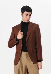 Rjav suknjič v teksturirani tkanini z dvema gumboma, odprtimi reverji in žepom na prsih, oblečen čez črn pulover s turtleneck izrezom.