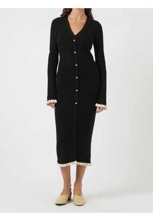 Femme portant une robe midi noire côtelée avec des poignets et un ourlet beiges à volants, boutonnée à l'avant, associée à des chaussures beiges à talons bas.