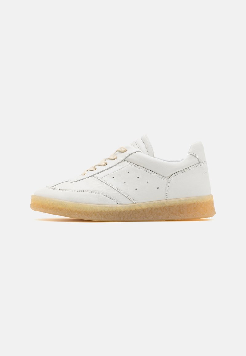 Baskets basses blanches avec des lacets beige, des détails perforés sur les côtés et une semelle en gomme marron clair sur fond uni.