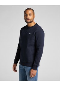 Mörkblå sweatshirt tillverkad av mjukt material, med rundad hals, ribbade ärmslut och bröstlogo. Kombinerad med ljusblå jeans.