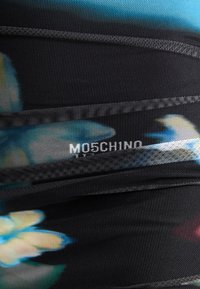 Črn materiaal z rožnatim vzorcem v modri in rumeni barvi, z belim logotipom "MOSCHINO" s teksturirano obdelavo in detajli šivanja.