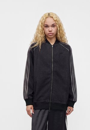 Junge Person mit langen lockigen blonden Haaren, die eine schwarze Jacke mit grauen Streifen an den Ärmeln trägt, vor einem schlichten weißen Hintergrund stehend.