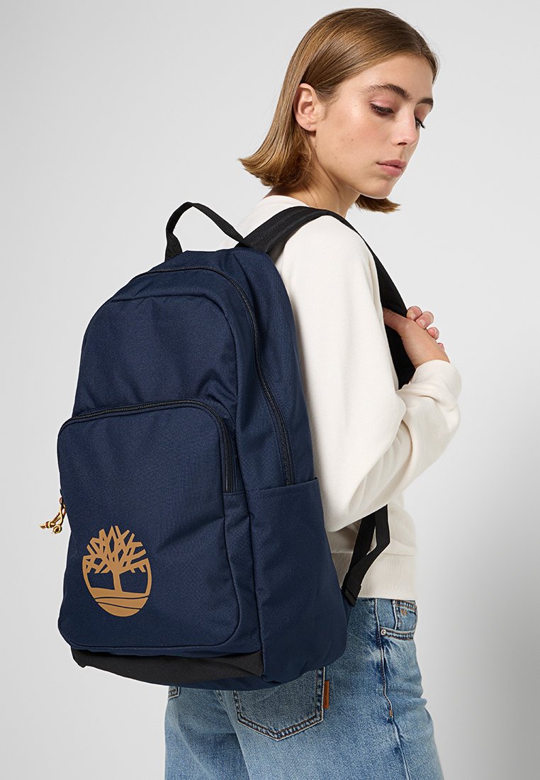 Sac à dos bleu marine en tissu, avec une poche zippée à l'avant et un logo d'arbre en brun bien visible. Bandoulières réglables.